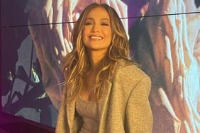 Jennifer Lopez Smiling Sitting Against Ledge Wearing Beige Coat and Matching Minidress