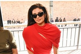 Kylie Jenner Sitting Red Dress Closed-Mouth Smile Hand on Leg Other Hand on Bench at "Les Sculptures" Jacquemus Fashion Show 