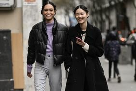A woman wears a black puffer jacket next to a woman wearing a long black wool coat, two types of coat to try for 2023.