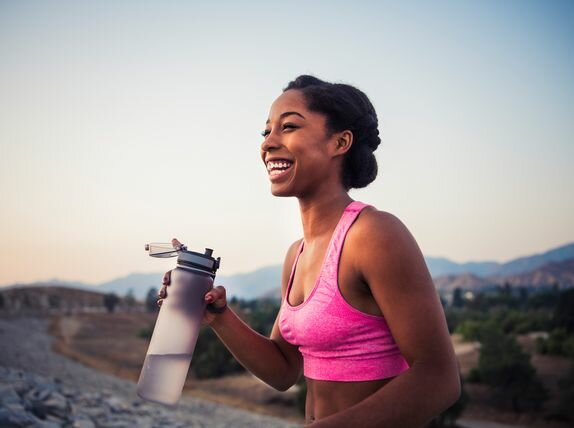 A smiling woman in workout clothing is standing outside holding a water bottle.