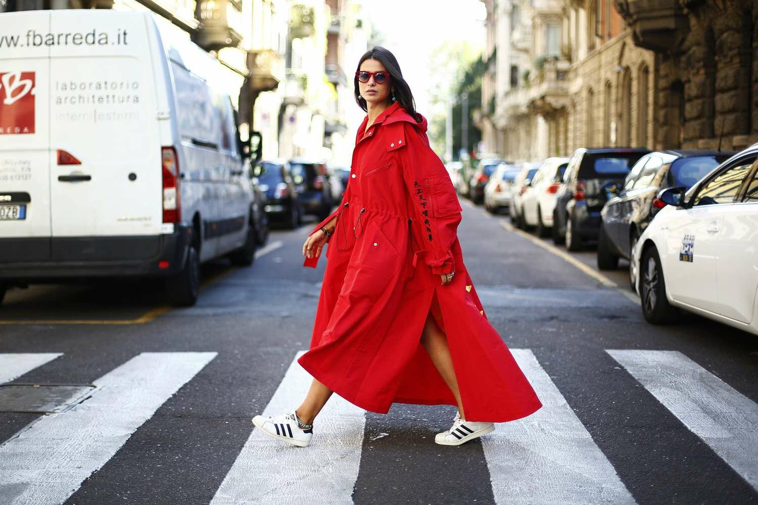 Woman walking across a city street in an oversized red trench coat and adidas court shoes
